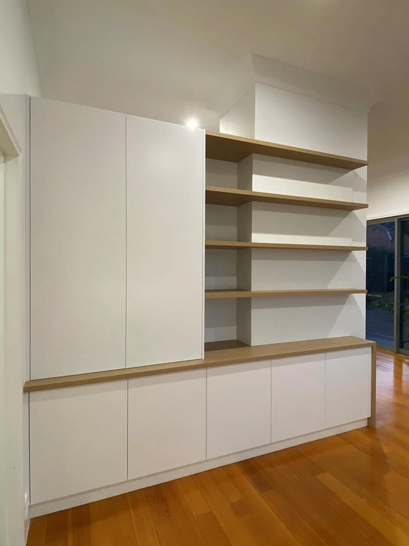 A spacious room with white walls and warm wooden floors, lined with shelves that hold books and decorative objects