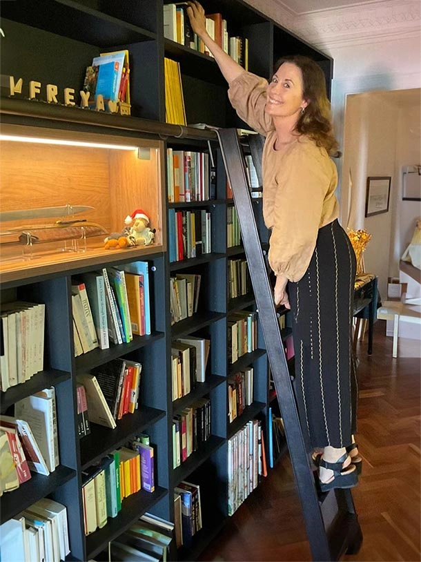 A woman on a ladder reaching for a book on a high shelf in a well-organized library setting