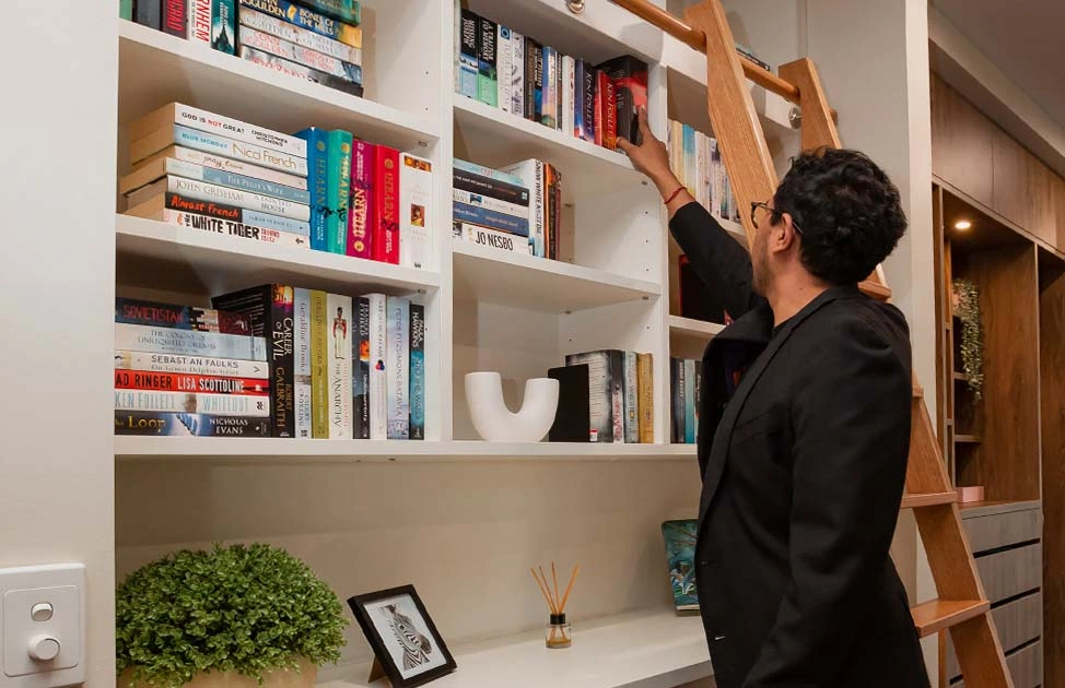 A man reaches upward, trying to take a book from a tall shelf stacked with numerous books of different sizes